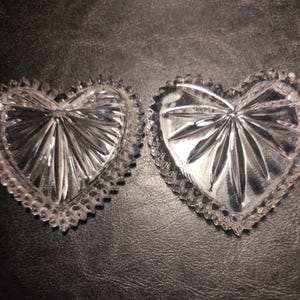 May include: Two clear, heart-shaped glass dishes with a starburst design in the center. The edges of the dishes have a scalloped, textured border. The dishes are displayed on a dark, textured surface.