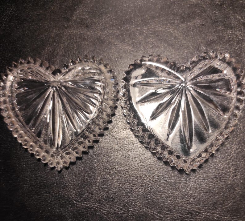May include: Two clear, heart-shaped glass dishes with a starburst design in the center. The edges of the dishes have a scalloped, textured border. The dishes are displayed on a dark, textured surface.