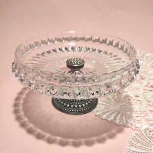 May include: A clear glass pedestal dish featuring a circular bowl with a scalloped rim and a radiating sunburst pattern on the base. The dish is supported by a dark, textured metal stand with a small, circular, beaded decorative accent.