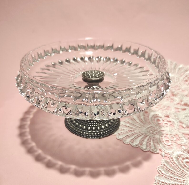 May include: A clear glass pedestal dish featuring a circular bowl with a scalloped rim and a radiating sunburst pattern on the base. The dish is supported by a dark, textured metal stand with a small, circular, beaded decorative accent.