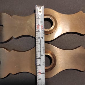 May include: Two brass-colored metal pieces with a decorative shape, each featuring a central circular opening. A measuring tape shows the pieces are approximately 4 inches in length. The background is a dark gray surface.