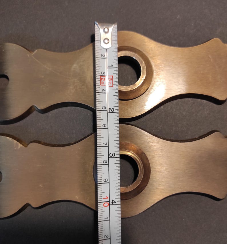 May include: Two brass-colored metal pieces with a decorative shape, each featuring a central circular opening. A measuring tape shows the pieces are approximately 4 inches in length. The background is a dark gray surface.