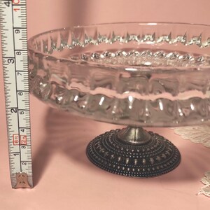 May include: A clear glass pedestal bowl with a textured rim and a dark, ornate metal base. A measuring tape placed vertically next to the bowl indicates a height of approximately 9 cm from the base to the top edge of the glass bowl.