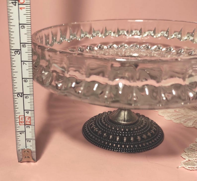 May include: A clear glass pedestal bowl with a textured rim and a dark, ornate metal base. A measuring tape placed vertically next to the bowl indicates a height of approximately 9 cm from the base to the top edge of the glass bowl.