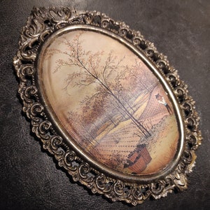 May include: An oval-shaped framed painting with a detailed ornate silver frame. The painting depicts a rural landscape with a tree in the center and a figure in the distance.