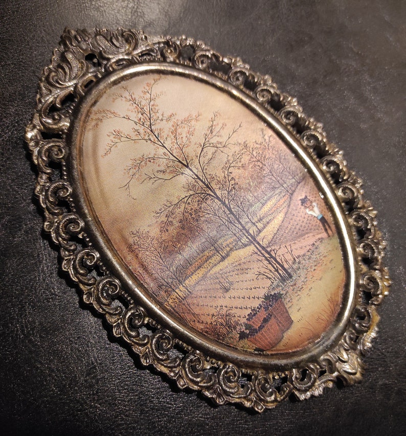 May include: An oval-shaped framed painting with a detailed ornate silver frame. The painting depicts a rural landscape with a tree in the center and a figure in the distance.
