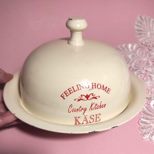 Può includere: Una campana per formaggio in smalto color crema con base abbinata e pomello. La campana presenta la scritta rossa "FEELING HOME Country Kitchen KÄSE" con un fregio decorativo. I bordi mostrano lievi segni di usura e metallo scuro.
