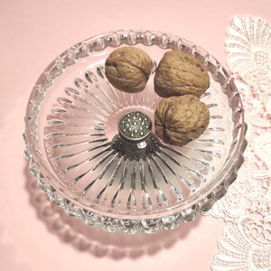 May include: A clear glass bowl with a radial ribbed pattern and a scalloped edge. The bowl features a central silver-toned metal post with a textured, circular top. Three whole brown walnuts are placed inside the glass dish.
