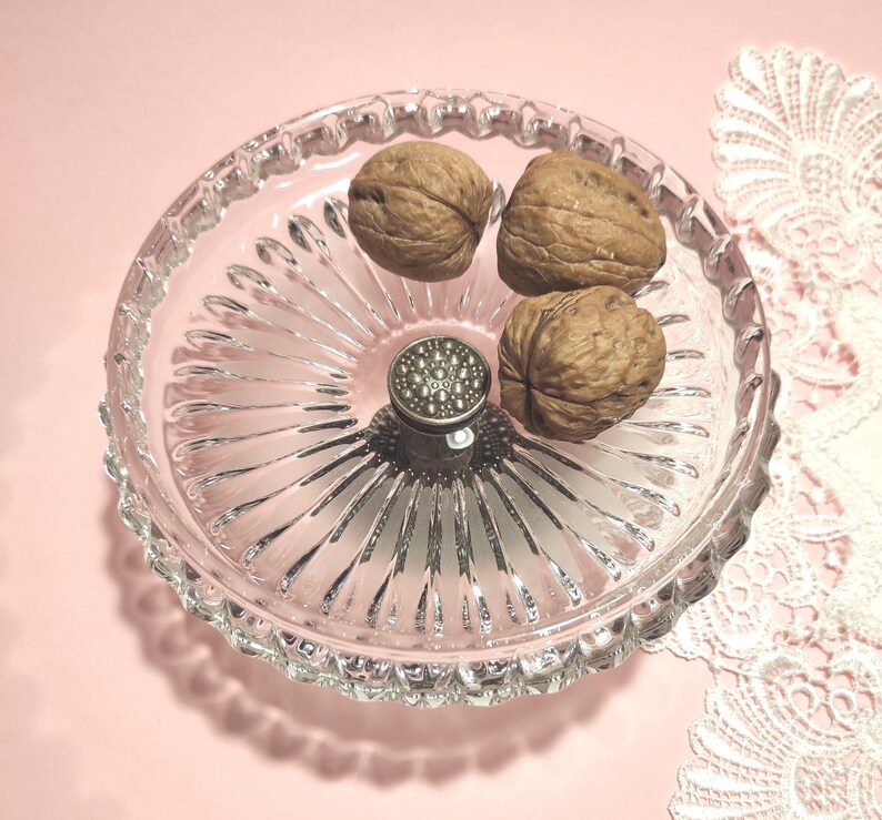 May include: A clear glass bowl with a radial ribbed pattern and a scalloped edge. The bowl features a central silver-toned metal post with a textured, circular top. Three whole brown walnuts are placed inside the glass dish.