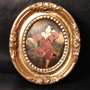 Può includere: Cornice ovale dorata con una stampa vintage di un bambino in abito rosso. La cornice presenta dettagli ornati e una finitura lucida. Il bambino è in posa dinamica su uno sfondo tenue.