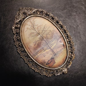May include: An oval-shaped framed painting with a gold-colored ornate frame. The painting depicts a landscape scene with trees and a figure in the distance.