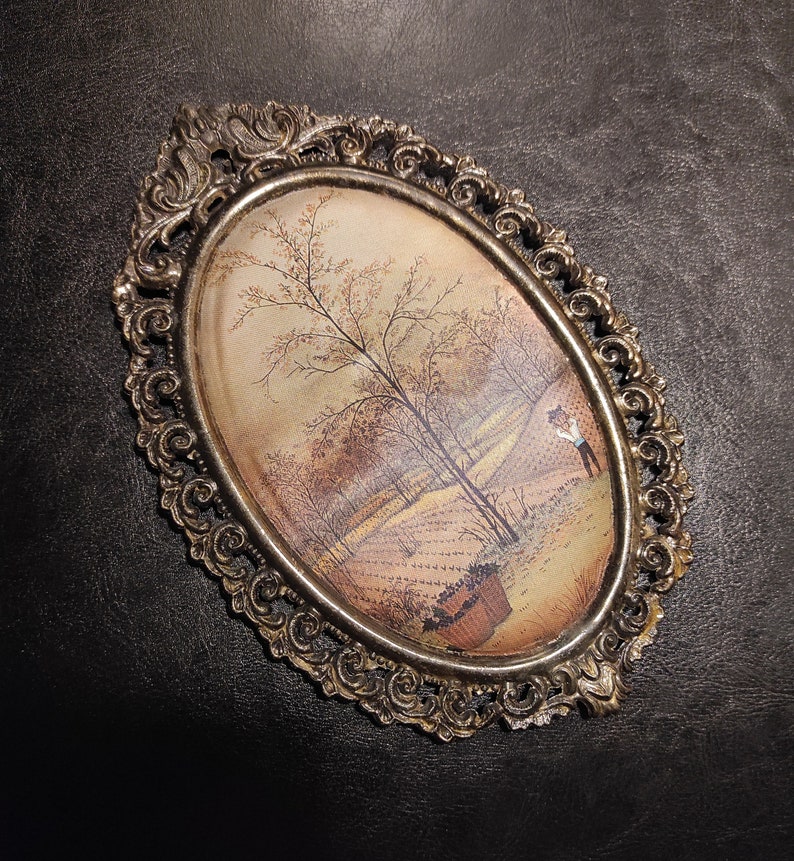May include: An oval-shaped framed painting with a gold-colored ornate frame. The painting depicts a landscape scene with trees and a figure in the distance.