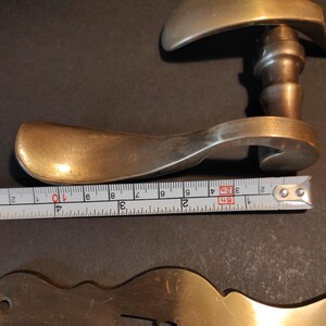 May include: Antique brass door handle with a curved design. The handle is approximately 4 inches long. A metal tape measure is placed next to the handle.