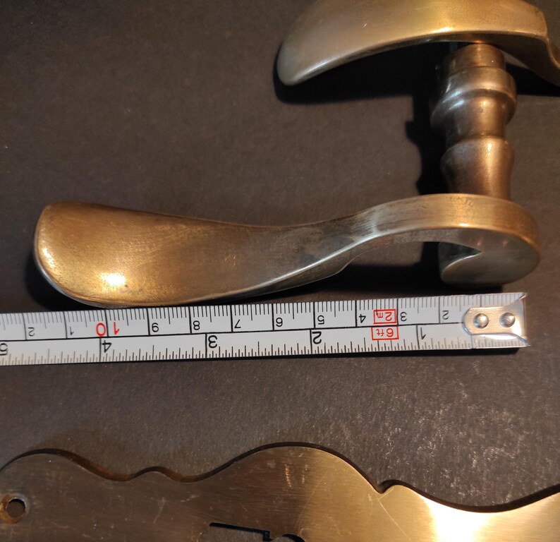 May include: Antique brass door handle with a curved design. The handle is approximately 4 inches long. A metal tape measure is placed next to the handle.