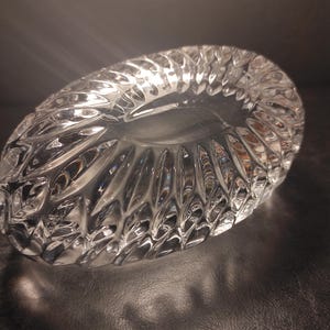 May include: A clear, oval-shaped crystal bowl with a textured, cut-glass design. The bowl's surface reflects light, creating a sparkling effect. The intricate pattern adds a touch of elegance to the decorative piece.