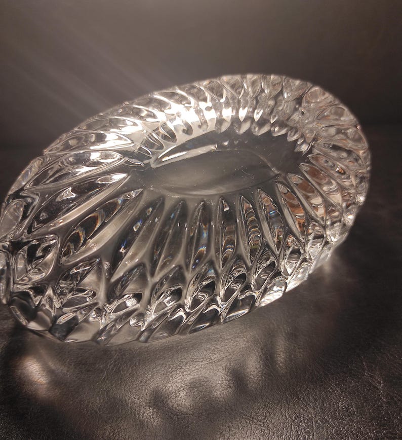 May include: A clear, oval-shaped crystal bowl with a textured, cut-glass design. The bowl's surface reflects light, creating a sparkling effect. The intricate pattern adds a touch of elegance to the decorative piece.