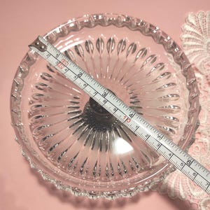 May include: A clear glass pedestal dish with a scalloped rim and radiating vertical ridges. A white measuring tape is placed across the diameter, showing a measurement of approximately 16.5 cm from edge to edge of the glass bowl.