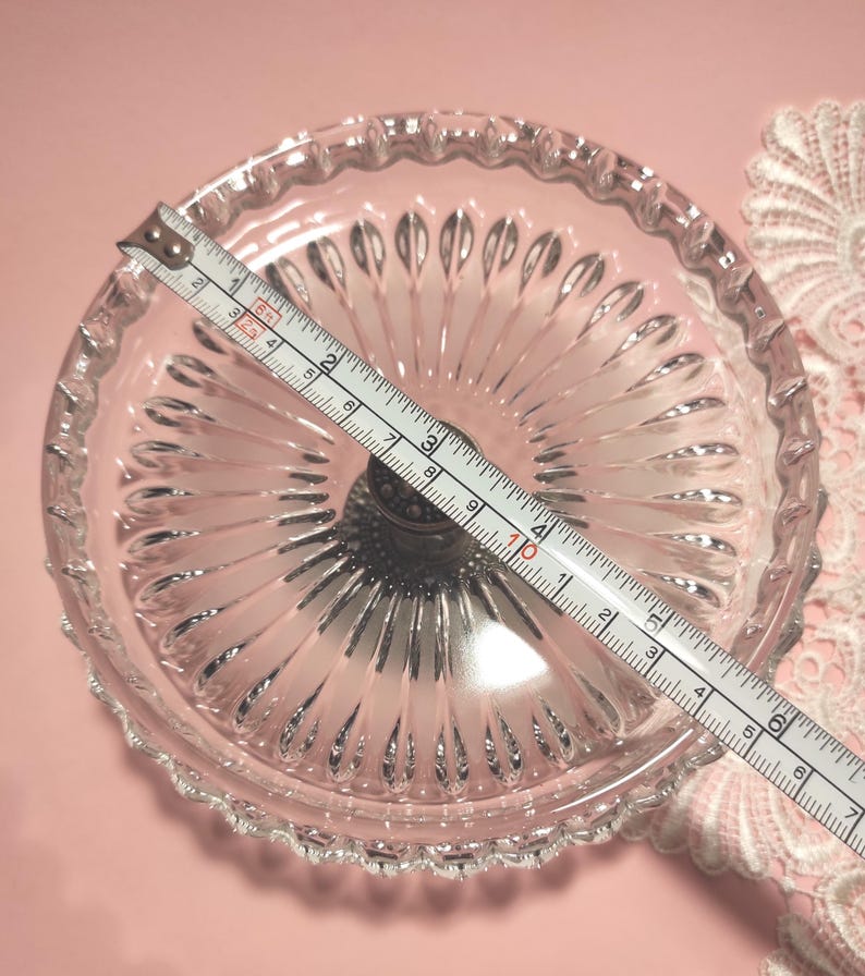 May include: A clear glass pedestal dish with a scalloped rim and radiating vertical ridges. A white measuring tape is placed across the diameter, showing a measurement of approximately 16.5 cm from edge to edge of the glass bowl.
