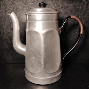 May include: A vintage aluminium coffee pot with a black handle and lid knob. The pot has a spout and a lid. The handle is black with a brown accent. The pot is silver and has a classic design.