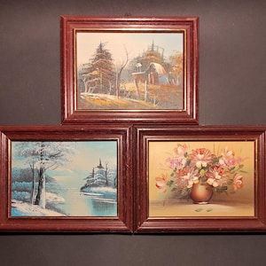 May include: Three framed paintings with different scenes. The top painting shows a rural landscape with a red barn and trees. The middle painting shows a snowy winter scene with a river and trees. The bottom painting shows a bouquet of flowers in a vase.