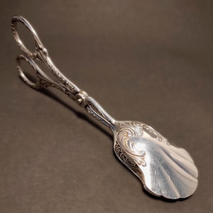 Op de afbeelding: Een zilverkleurige metalen serveertang met een gepolijste afwerking en gedetailleerde, in reliëf aangebrachte krulpatronen op de handvatten en de schep. Het keukengerei heeft twee ronde vingerringen en een breed, schelpvormig uiteinde.