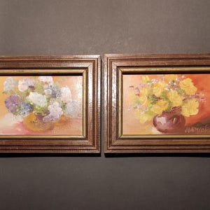 May include: Two framed oil paintings of floral arrangements. The painting on the left features a bouquet of purple and white flowers in a brown vase. The painting on the right features a bouquet of yellow flowers in a purple vase. Both paintings have a warm, orange background.