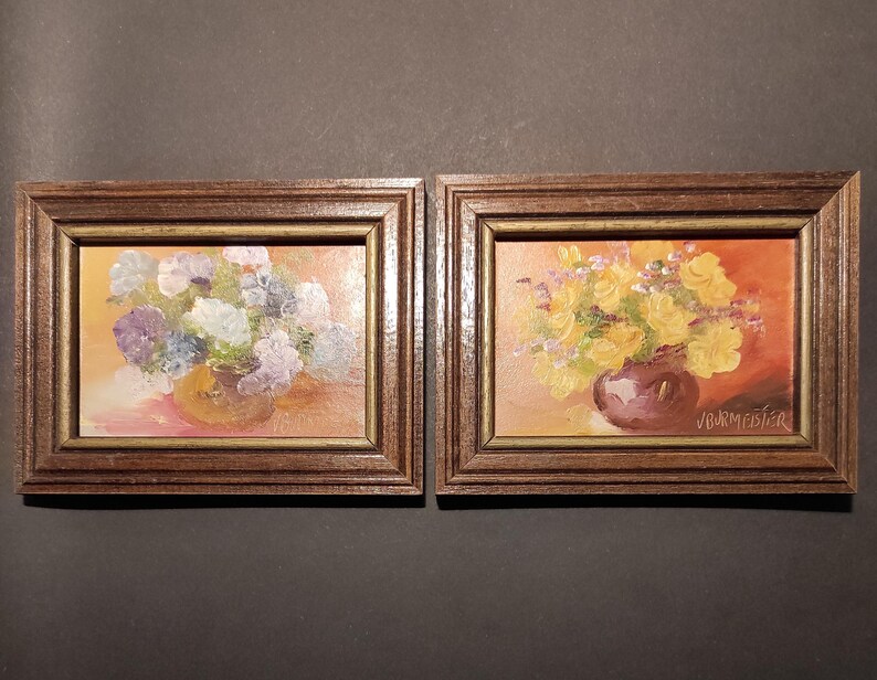 May include: Two framed oil paintings of floral arrangements. The painting on the left features a bouquet of purple and white flowers in a brown vase. The painting on the right features a bouquet of yellow flowers in a purple vase. Both paintings have a warm, orange background.