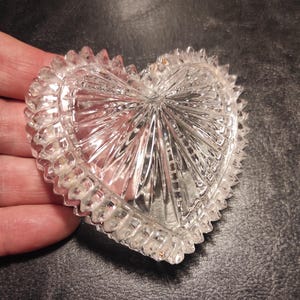 May include: Clear, heart-shaped glass dish with a textured, starburst design. The rim features a series of small, pointed edges. The dish is held in a hand, showcasing its intricate details and reflective qualities.