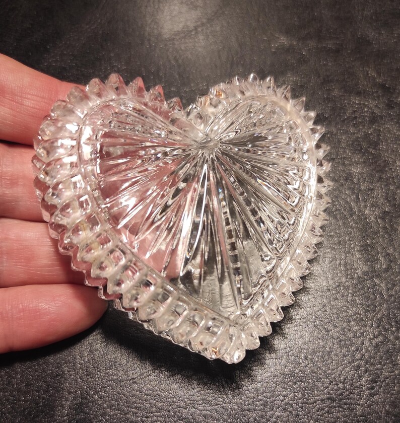 May include: Clear, heart-shaped glass dish with a textured, starburst design. The rim features a series of small, pointed edges. The dish is held in a hand, showcasing its intricate details and reflective qualities.