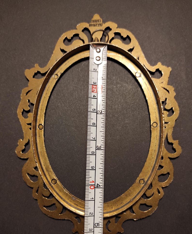 Puede incluir: Marco de fotos ovalado de lat&oacute;n con adornos decorativos. La altura interior del marco es de aproximadamente 10 cm, como se muestra en una cinta m&eacute;trica. El marco est&aacute; marcado "MADE IN ITALY".