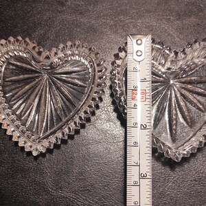 May include: Two clear, heart-shaped glass dishes with a starburst design in the center and a scalloped edge. A ruler shows the dishes are approximately 3 inches wide. The dishes are on a dark surface.