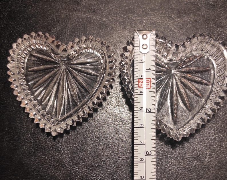May include: Two clear, heart-shaped glass dishes with a starburst design in the center and a scalloped edge. A ruler shows the dishes are approximately 3 inches wide. The dishes are on a dark surface.