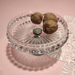 May include: A clear glass pedestal dish with a scalloped rim and a radiating sunburst pattern on the base. The dish features a central silver-toned metal accent with a textured surface and contains three whole brown walnuts inside the bowl.