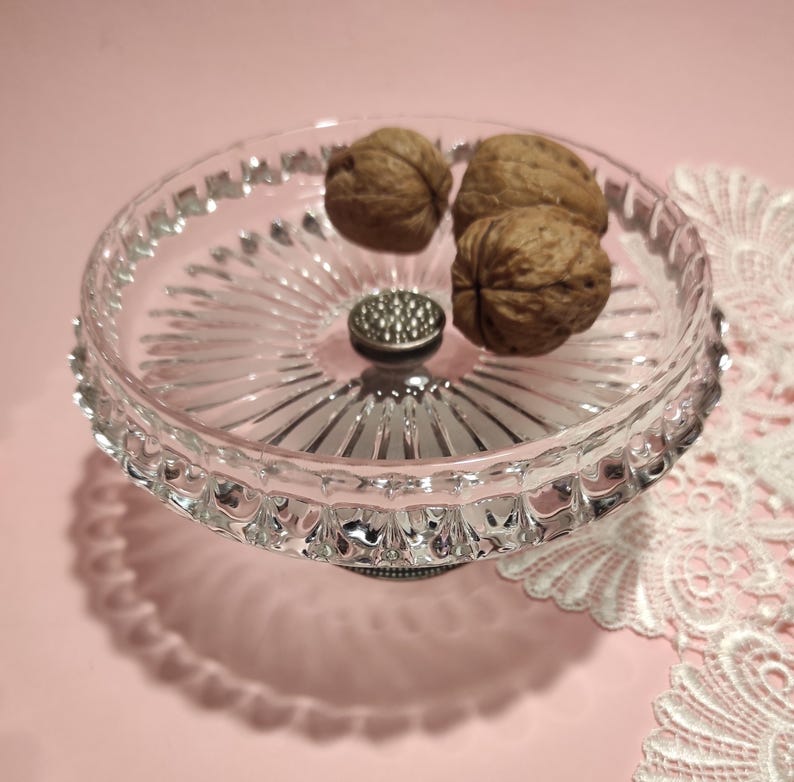 May include: A clear glass pedestal dish with a scalloped rim and a radiating sunburst pattern on the base. The dish features a central silver-toned metal accent with a textured surface and contains three whole brown walnuts inside the bowl.