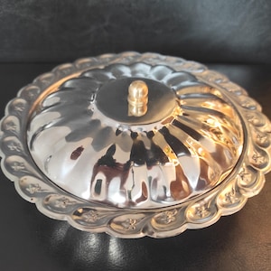 May include: A silver-coloured, round serving dish with a lid. The dish has a scalloped edge with floral detailing and a central knob on the lid. The reflective surface of the dish creates a bright, eye-catching appearance.