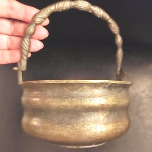 May include: A brass bucket with a rounded, ribbed body and a textured, decorative handle. The metal surface shows a weathered, aged patina with variations in color, featuring a wide opening and a curved base structure.
