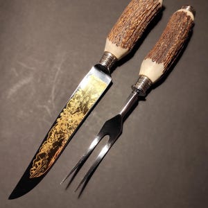 Op de afbeelding: Een vlees snijset met een mes en een vork. Het mes heeft een zwart lemmet met gouden filigraan en een bruine geweihandgreep. De vork is van roestvrij staal met een bijpassende geweihandgreep.