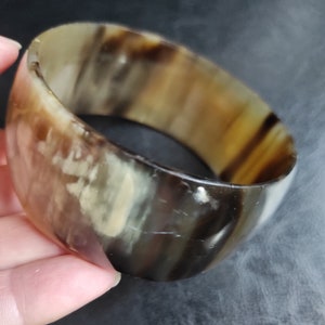 Könnte beinhalten: Ein breites, braunes und cremefarbenes Hornarmband. Das Armband ist aus poliertem Horn gefertigt und hat ein natürliches, organisches Aussehen.