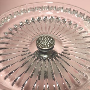 May include: A clear glass serving dish featuring a radial sunburst pattern of pressed glass ridges extending from the centre. The centre of the dish is topped with a small, circular, textured silver-toned metal knob with a dotted surface.