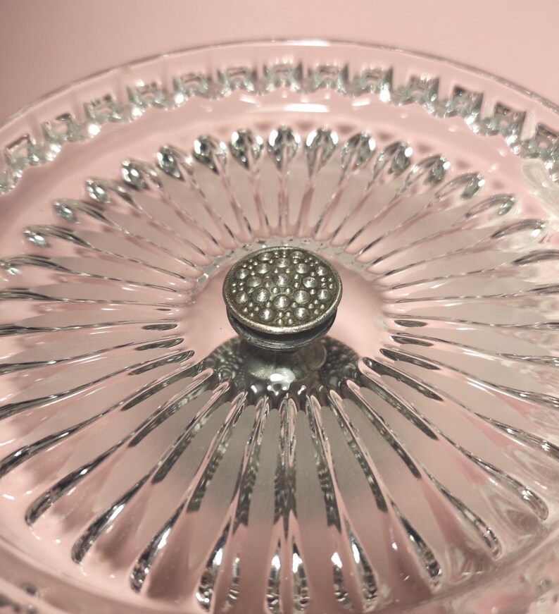 May include: A clear glass serving dish featuring a radial sunburst pattern of pressed glass ridges extending from the centre. The centre of the dish is topped with a small, circular, textured silver-toned metal knob with a dotted surface.