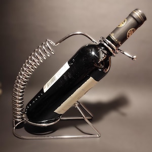 Puede incluir: Un porta botellas de vino de metal plateado con un brazo de resorte y una base triangular. Una botella de vino tinto descansa en el soporte.
