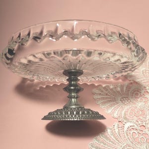 May include: A clear glass pedestal dish featuring a scalloped rim and a radial pattern on the base. The dish is supported by a metallic, silver-toned stem with a textured, beaded circular base, providing a stable foundation for the glass bowl.