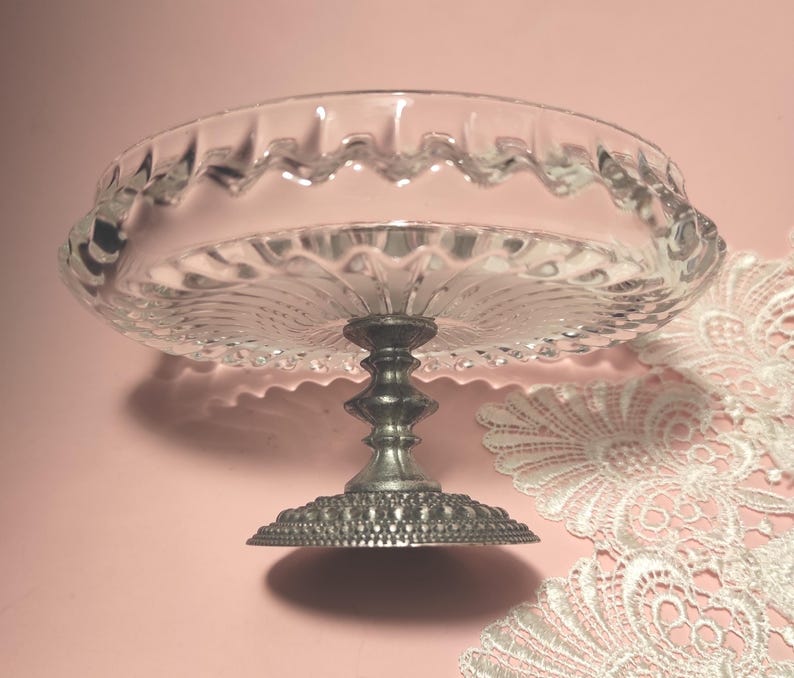 May include: A clear glass pedestal dish featuring a scalloped rim and a radial pattern on the base. The dish is supported by a metallic, silver-toned stem with a textured, beaded circular base, providing a stable foundation for the glass bowl.