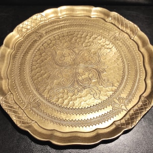 May include: A round gold-coloured metal tray with a scalloped edge. The surface features a central embossed floral scroll pattern surrounded by a textured hammered background, a decorative border, and a cross-hatched pattern along the rim.