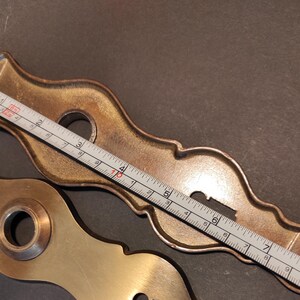 May include: Two antique brass door hardware pieces. One is a decorative escutcheon plate, approximately 8 inches long, with a keyhole and mounting holes. The other is a matching brass piece. A ruler is placed across the escutcheon plate.