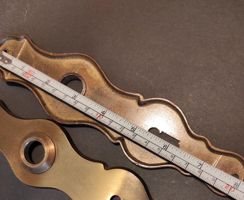 May include: Two antique brass door hardware pieces. One is a decorative escutcheon plate, approximately 8 inches long, with a keyhole and mounting holes. The other is a matching brass piece. A ruler is placed across the escutcheon plate.