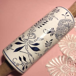 Può includere: Un mattarello bianco con un motivo floreale blu e manici in legno. Il mattarello in ceramica presenta un motivo ripetuto di fiori e foglie. Il mattarello è lungo circa 30 cm.