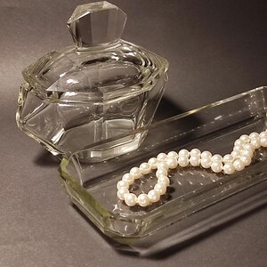 May include: A clear glass trinket dish with a string of white pearls inside. The dish has a rectangular shape with rounded corners.  The dish is sitting on a grey surface.