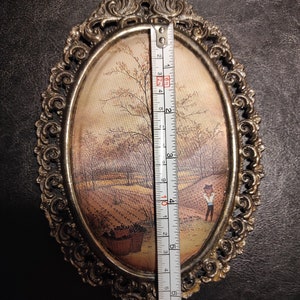 May include: An oval-shaped framed painting with a gold-colored metal frame. The painting depicts a rural scene with a small figure in the foreground. The frame has a decorative design.