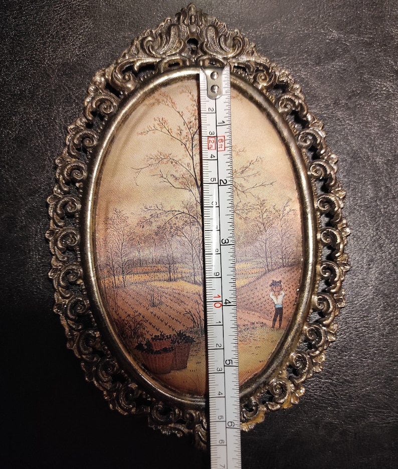May include: An oval-shaped framed painting with a gold-colored metal frame. The painting depicts a rural scene with a small figure in the foreground. The frame has a decorative design.